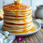 A stack of fluffy lemon poppy seed pancakes topped with a slice of lemon and a drizzle of syrup.